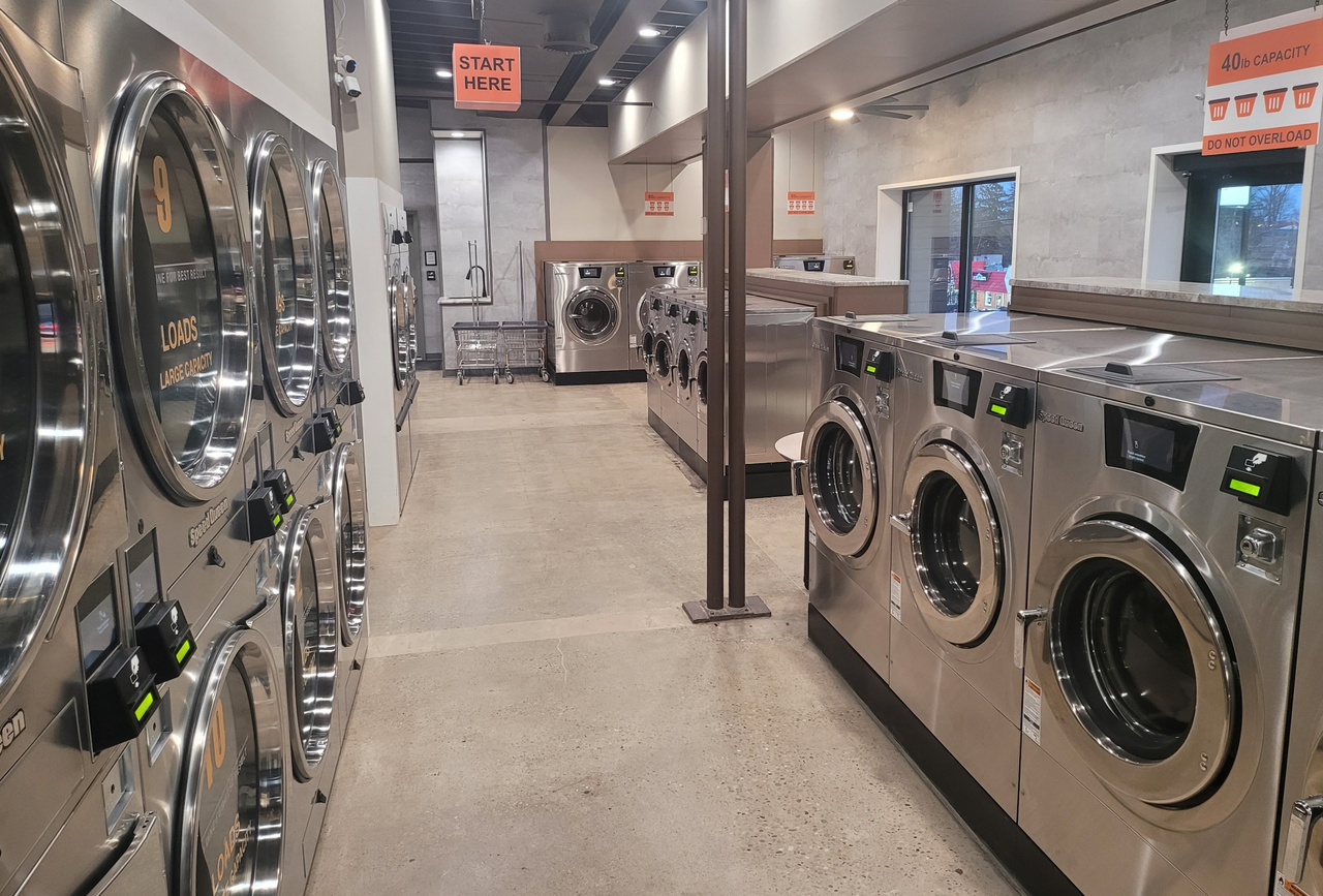 A long laundromat aisle with stainless washers and a start-here sign