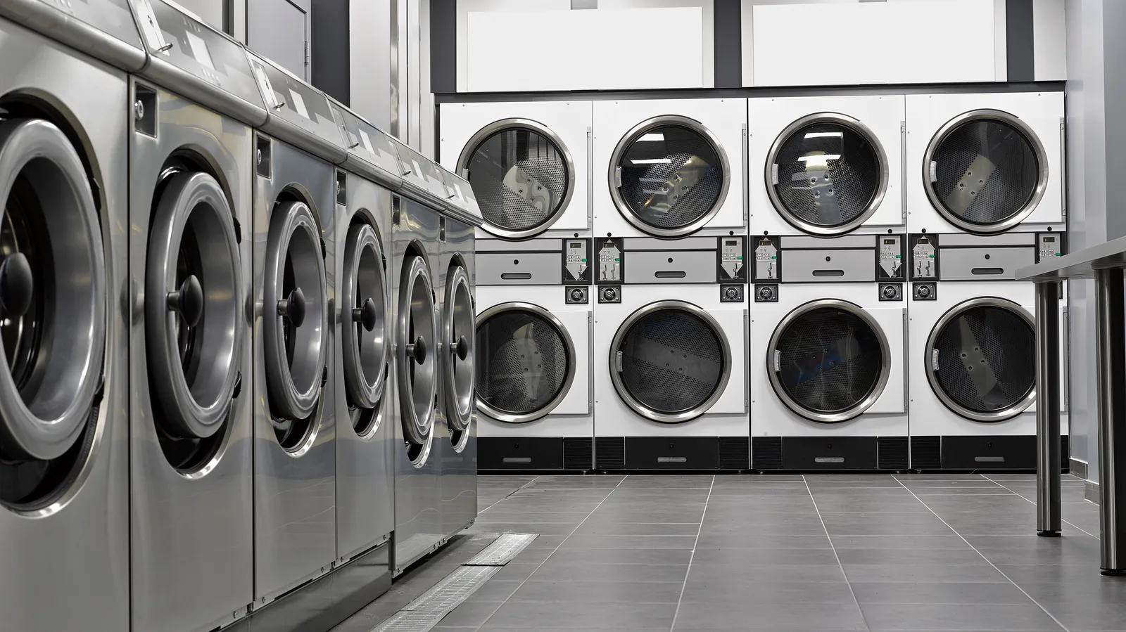 Stainless front-load washers lining a self-service laundry with stacked dryers along the back wall