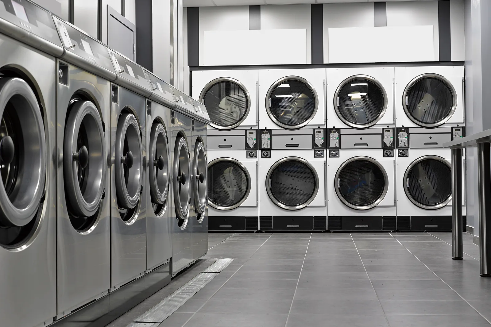 A self-service laundry with stainless washers down the left side and stacked dryers facing them across a tiled floor
