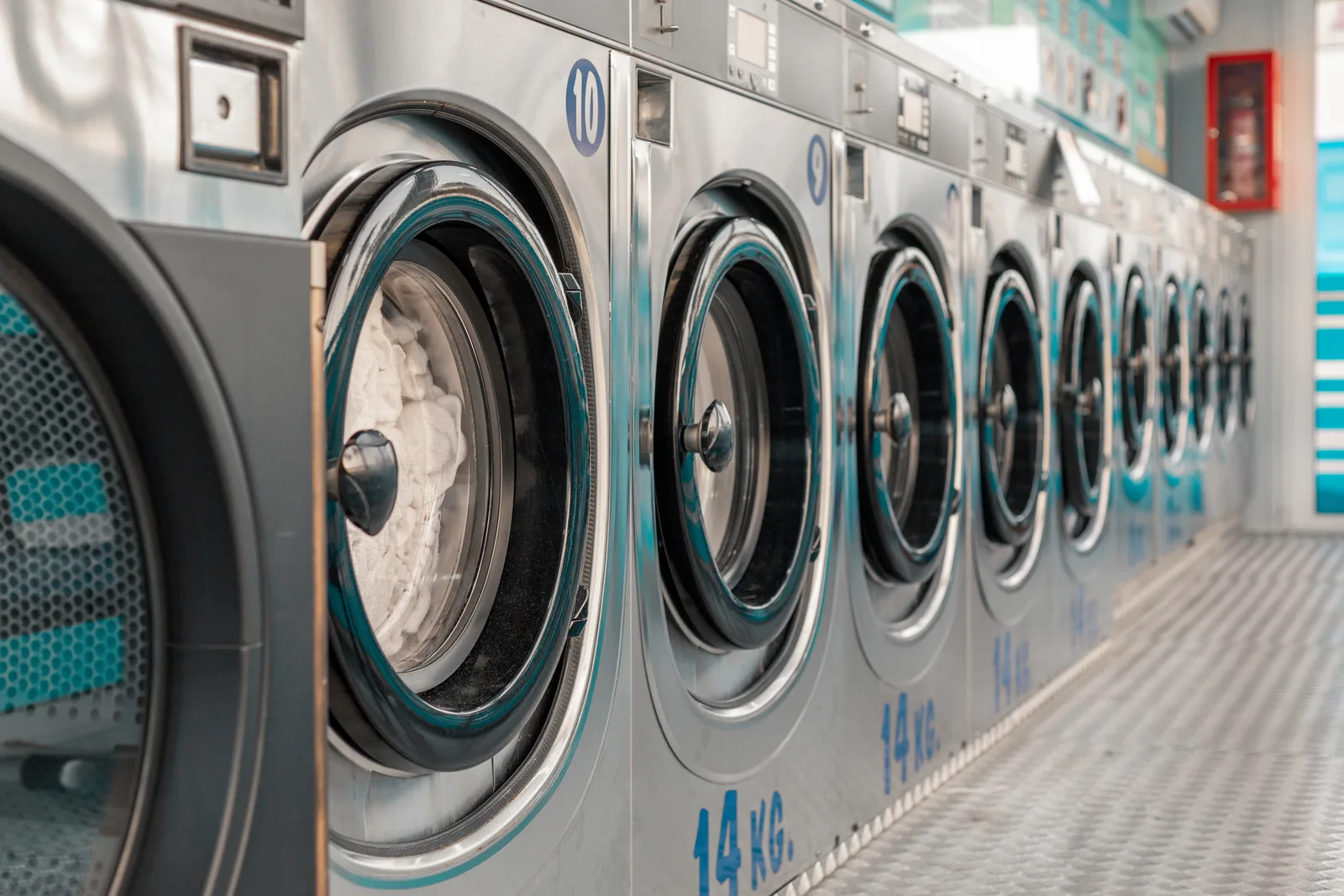 Front-load washers with trimmed door rings receding along a self-service laundry aisle, the nearest drum open and loaded