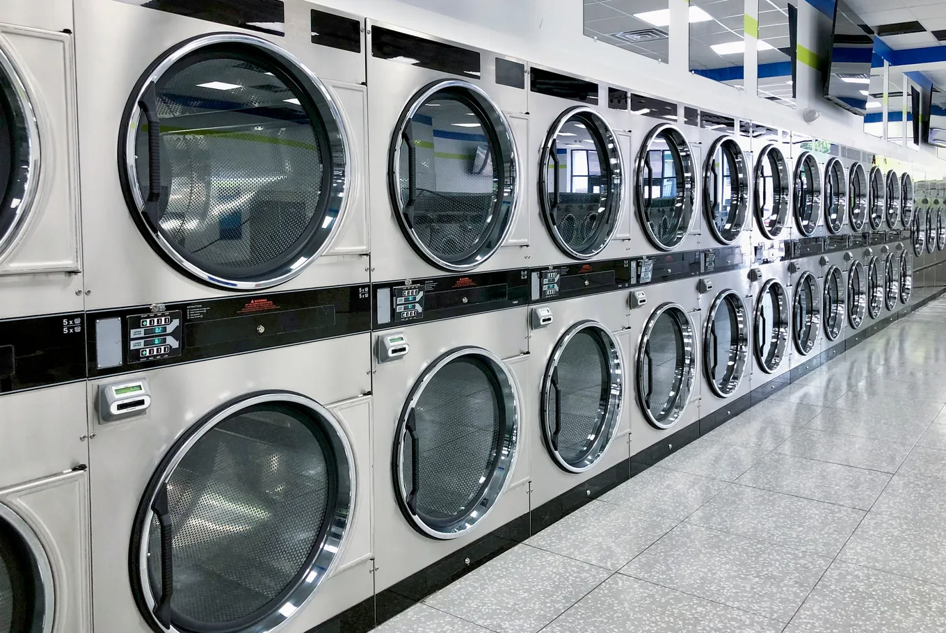 Stacked dryers with round chrome doors lining a laundry aisle under bright ceiling panels
